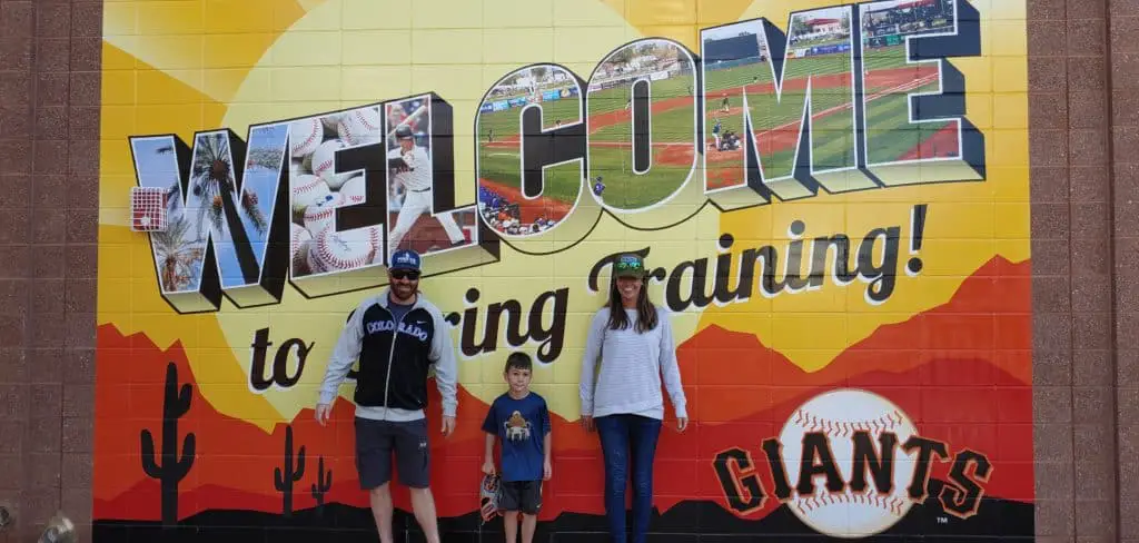Parents and a child standing in front of a Giants Spring Training Sign painted on a brick wall