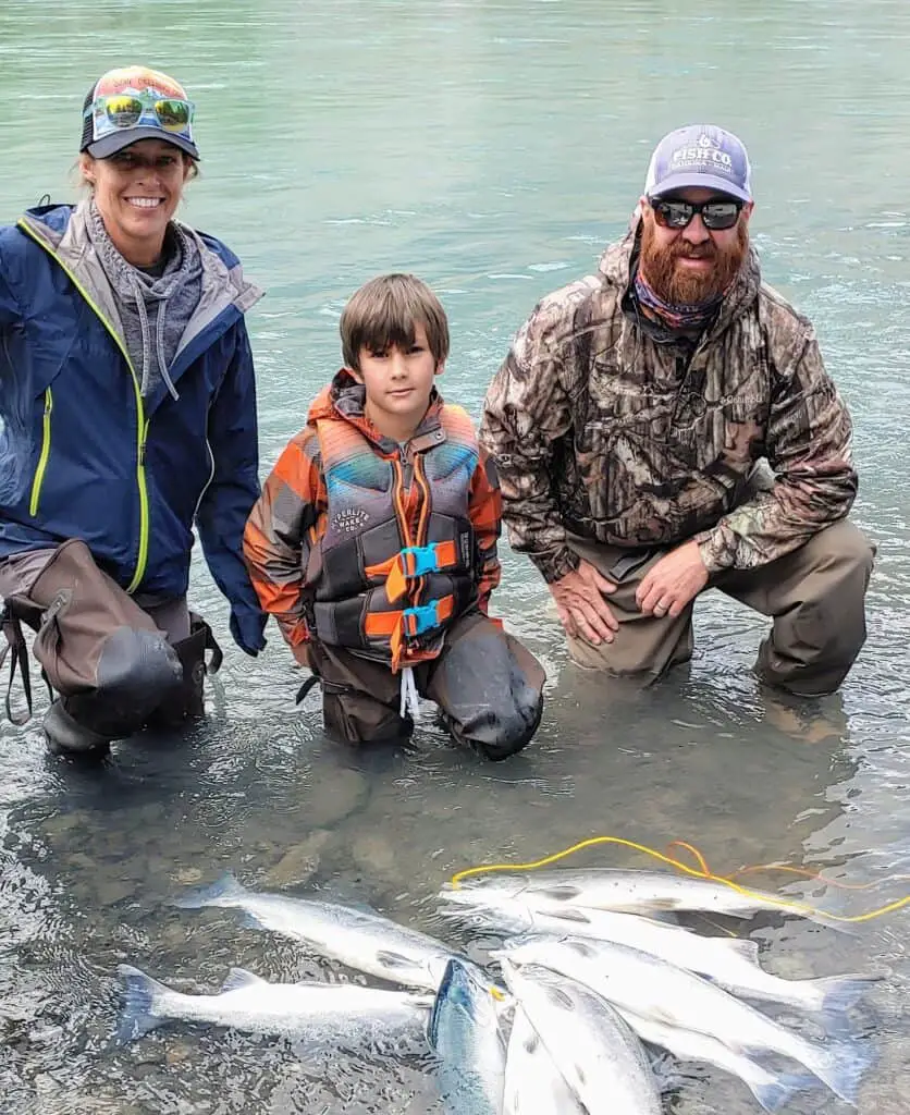 mom and dad and son in the river with all of the salmon they caught fishing in alaska in front of them