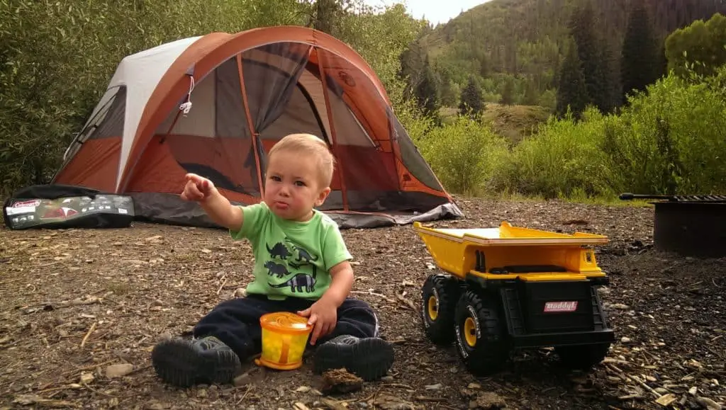 baby sitting in the dirt in front of a tent eating snacks,