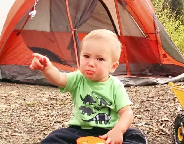 baby sitting in the dirt in front of a tent eating snacks