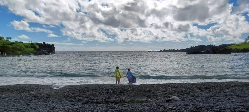 two boys with boogie boards looking out at the ocean from the black sand beach of Wai'anapanapa State Park